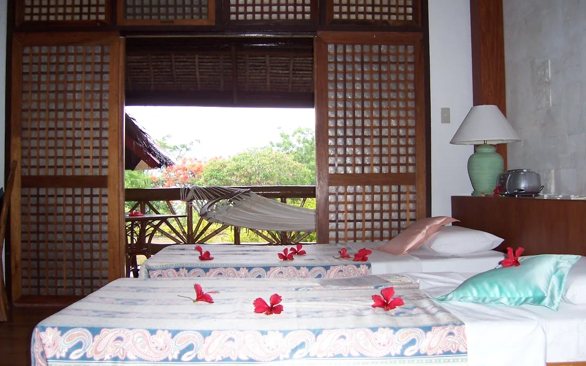 Photo: Bing Ramos / CC BY 2.0, via Wikimedia Commons A resort room on Badian’s Zaragosa Island, capiz shutters open onto a balcony hammock and flame tree, hibiscus flowers laid on the beds