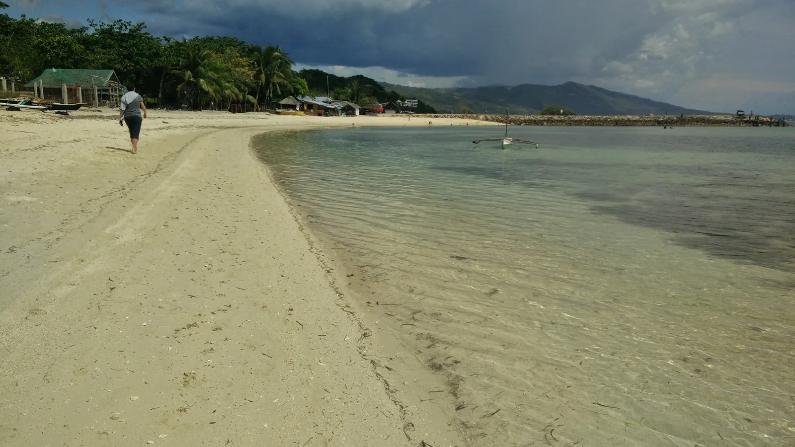 The long curve of white sand and shallow clear water at Lambug Beach, Badian, Cebu, with an outrigger moored offshore and mountains behind
