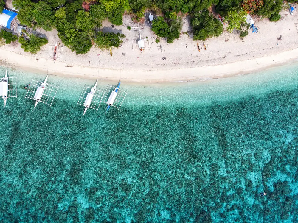 Aerial view of outrigger boats moored over clear turquoise water beside a white-sand beach